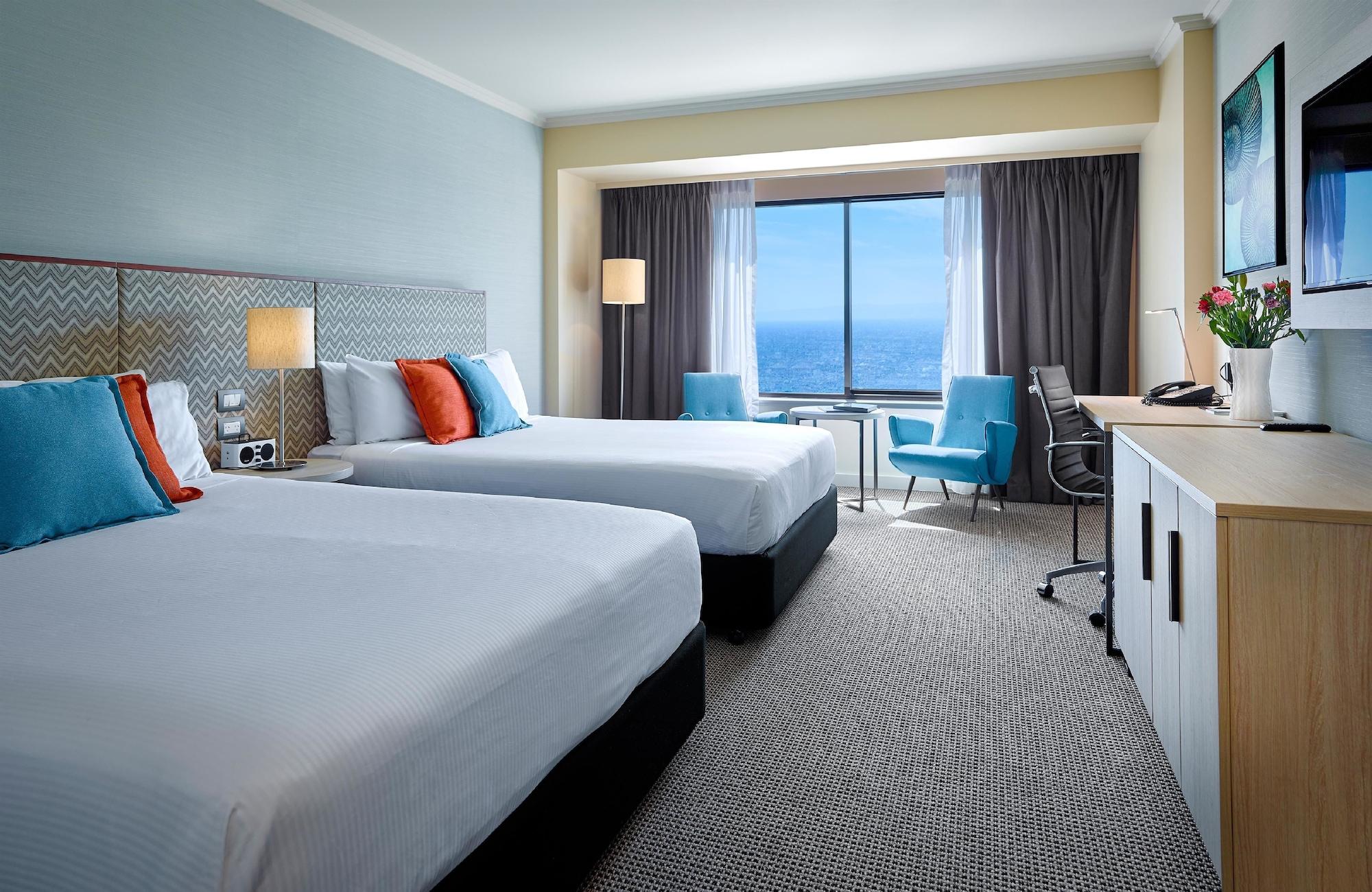 Stamford Grand Adelaide Hotel Glenelg Ngoại thất bức ảnh The photo shows a well-decorated hotel room featuring two queen-sized beds with white linens and colorful accent pillows. There is a large window with curtains that offers a view of the ocean. A small seating area with a blue chair and a matching des