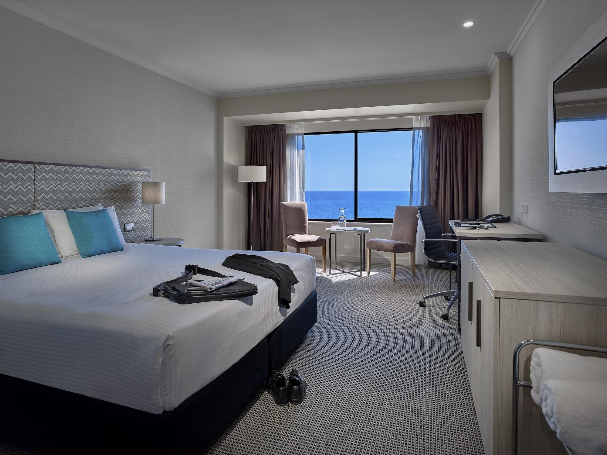 Stamford Grand Adelaide Hotel Glenelg Ngoại thất bức ảnh The photo shows a modern hotel room featuring a queen-sized bed with white linens and two aqua blue decorative pillows. There are bedside lamps and a large window with a view of the ocean. The room also has a small seating area with two chairs and a 
