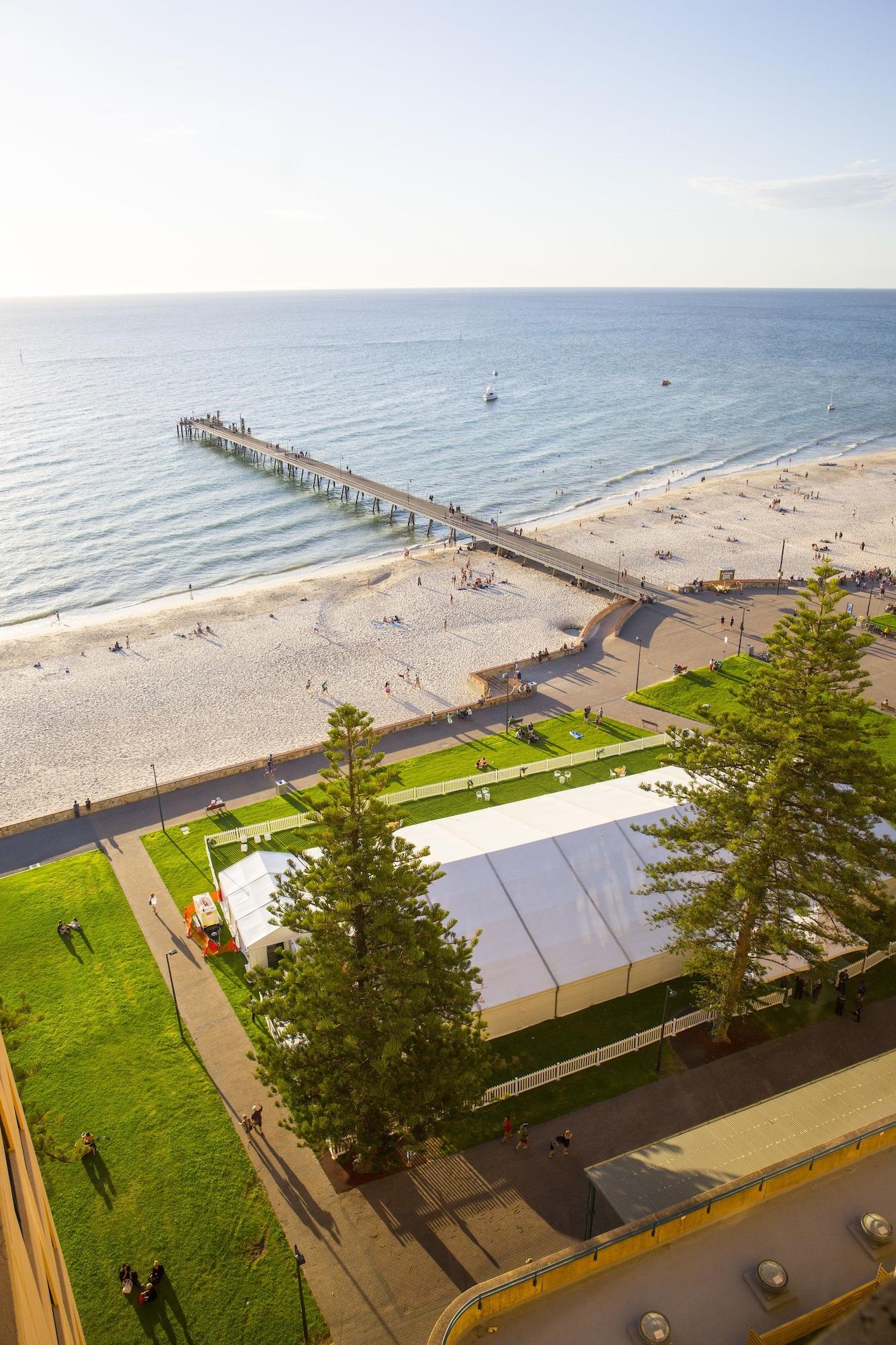 Stamford Grand Adelaide Hotel Glenelg Ngoại thất bức ảnh The photo shows a scenic view of a beach with a long pier extending into the water. The beach is sandy, and there are people enjoying the sun and walking along the shore. In the foreground, there is a large white tent set up on a grassy area, likely 