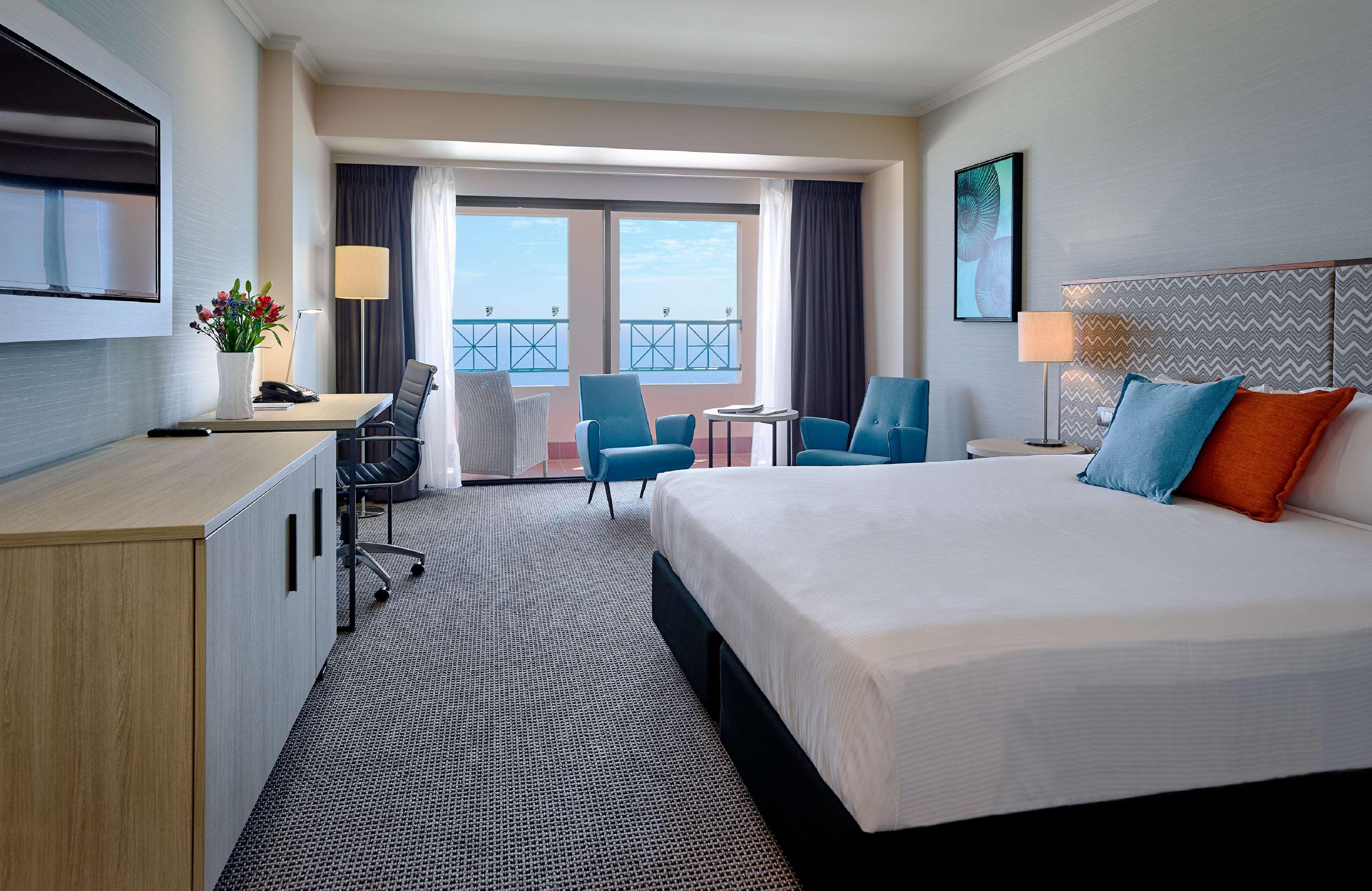 Stamford Grand Adelaide Hotel Glenelg Ngoại thất bức ảnh The photo depicts a modern hotel room. The room features a large bed with a white comforter, and there are decorative pillows in orange and blue. There is a flat-screen TV mounted on the wall, and a desk with an office chair is positioned near the wi