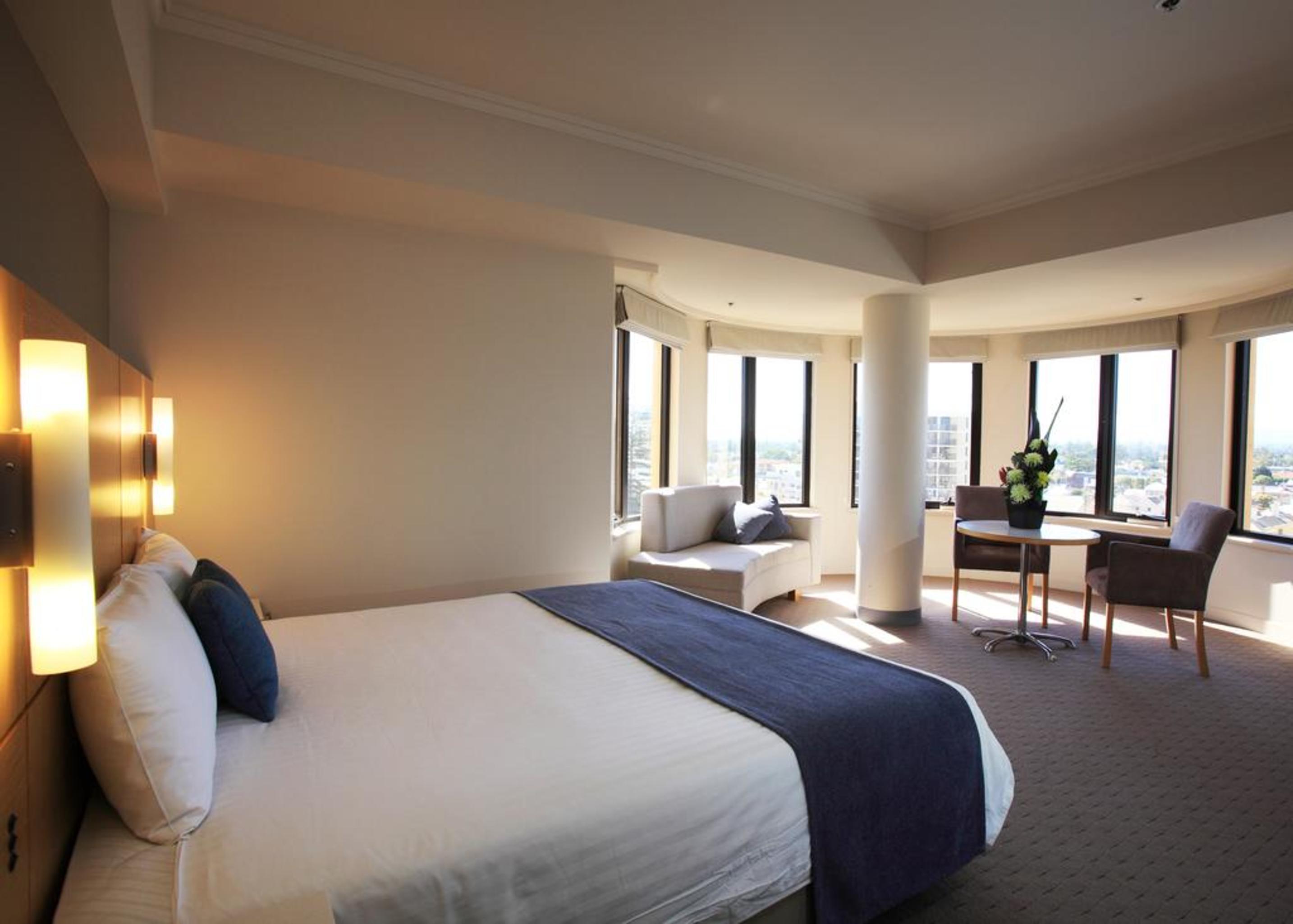 Stamford Grand Adelaide Hotel Glenelg Ngoại thất bức ảnh The photo shows a hotel room with a modern and spacious design. There is a large bed with white linens and a dark blue throw blanket. The room features multiple windows that allow plenty of natural light, and there is a small seating area near the wi