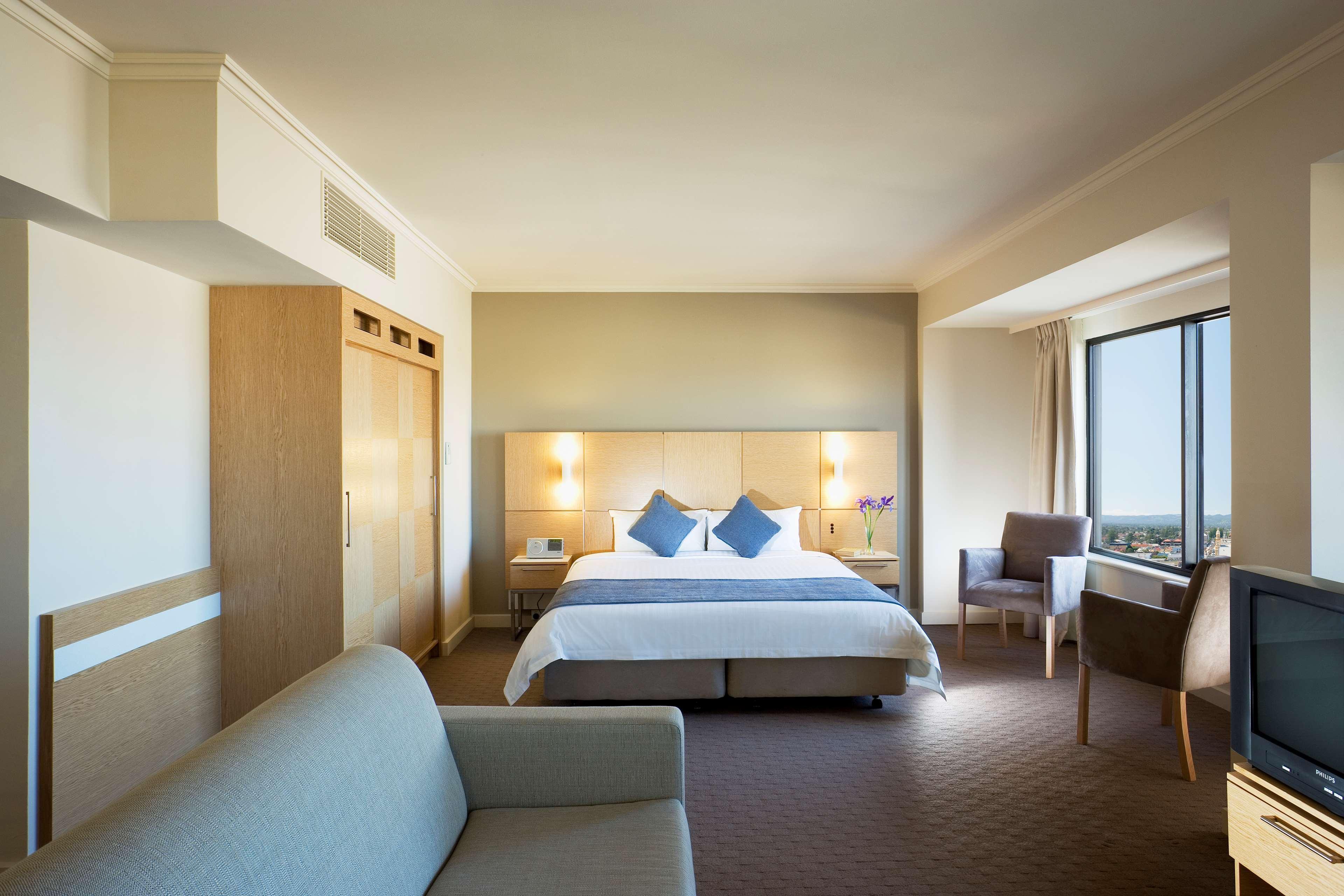 Stamford Grand Adelaide Hotel Glenelg Phòng bức ảnh The photo shows a modern hotel room featuring a large bed with two blue decorative pillows. There are bedside lamps on both sides of the headboard. To the left, there's a wooden wardrobe and a small table with a vase and a few amenities. On the right