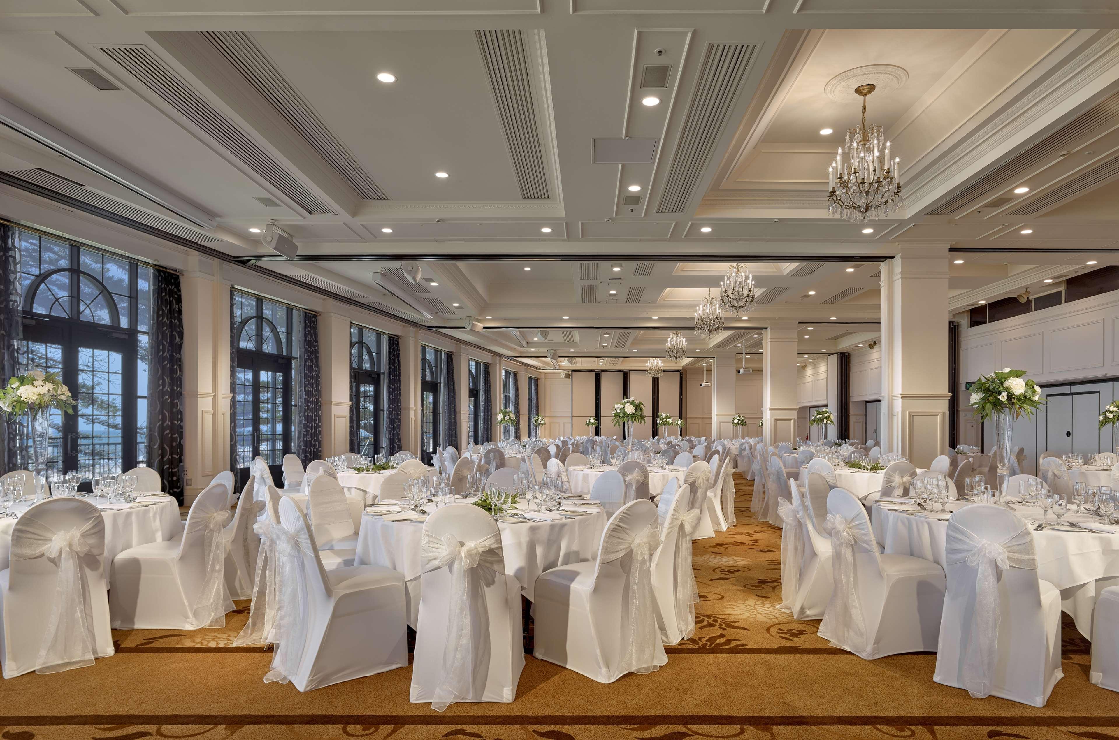 Stamford Grand Adelaide Hotel Glenelg Ngoại thất bức ảnh The photo depicts an elegantly arranged banquet hall or dining area. The space features multiple round tables, each set with white tablecloths and decorated with floral centerpieces. The chairs are covered in white fabric, contributing to a formal at