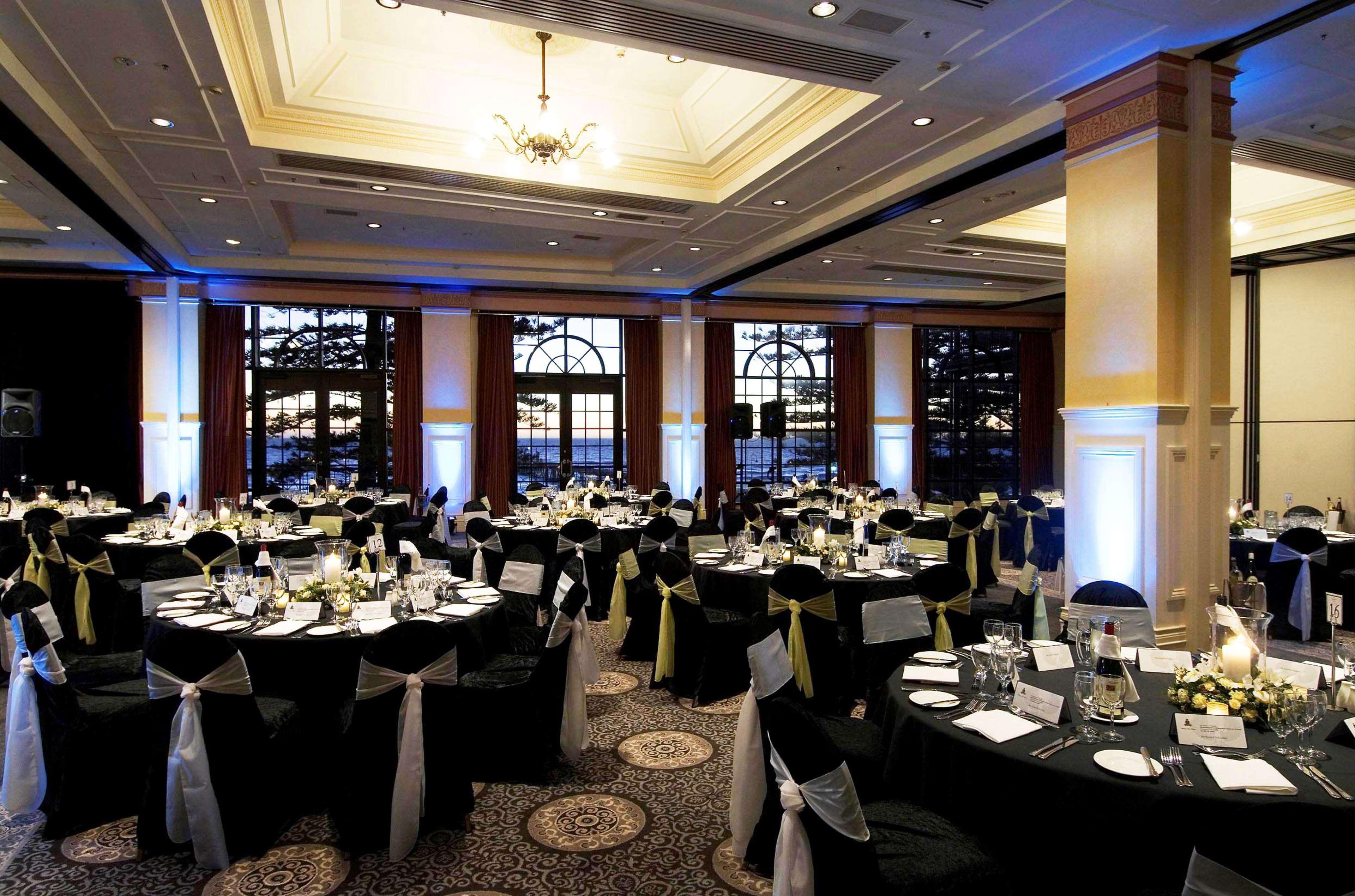 Stamford Grand Adelaide Hotel Glenelg Nhà hàng bức ảnh The photo depicts an elegantly arranged banquet hall set for an event. The room features round tables covered with black tablecloths, each adorned with white tableware and floral centerpieces. Chairs are draped in white fabric with decorative sashes 
