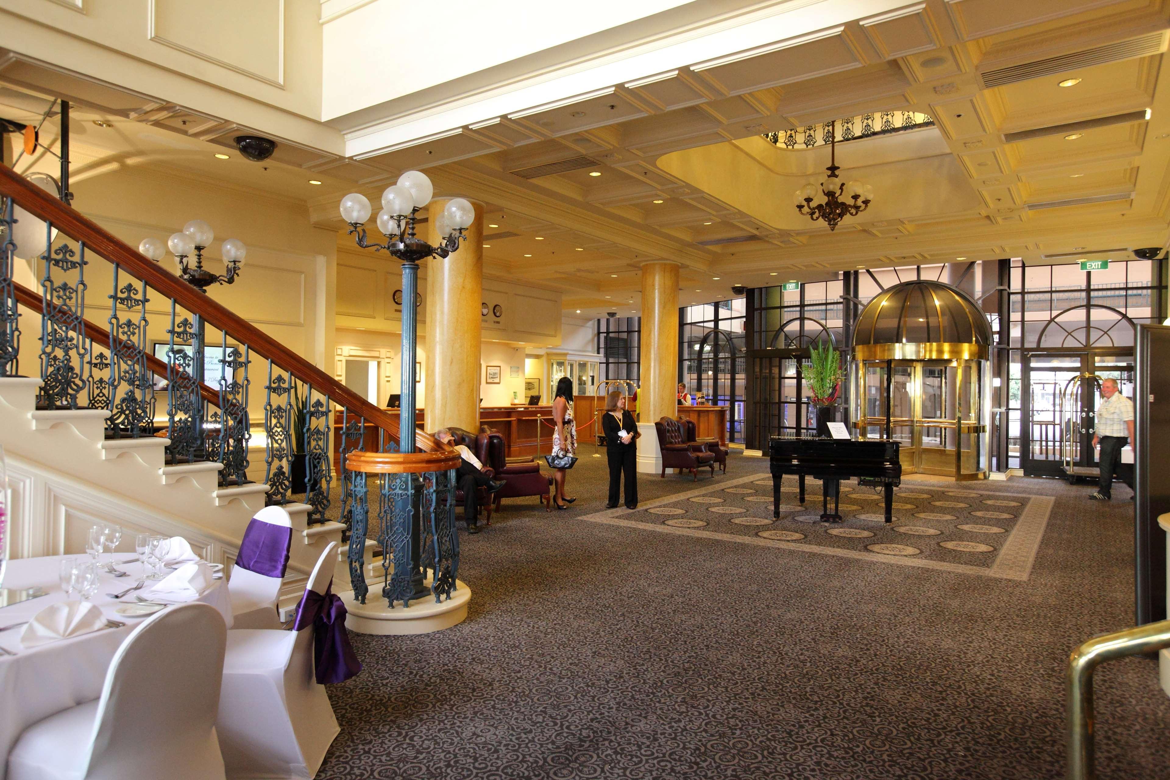 Stamford Grand Adelaide Hotel Glenelg Nội địa bức ảnh The photo shows a luxurious interior of a hotel or a restaurant lobby. There is an elegant staircase with ornate railings leading to an upper level. The space features high ceilings with decorative chandeliers and ample natural light coming through l