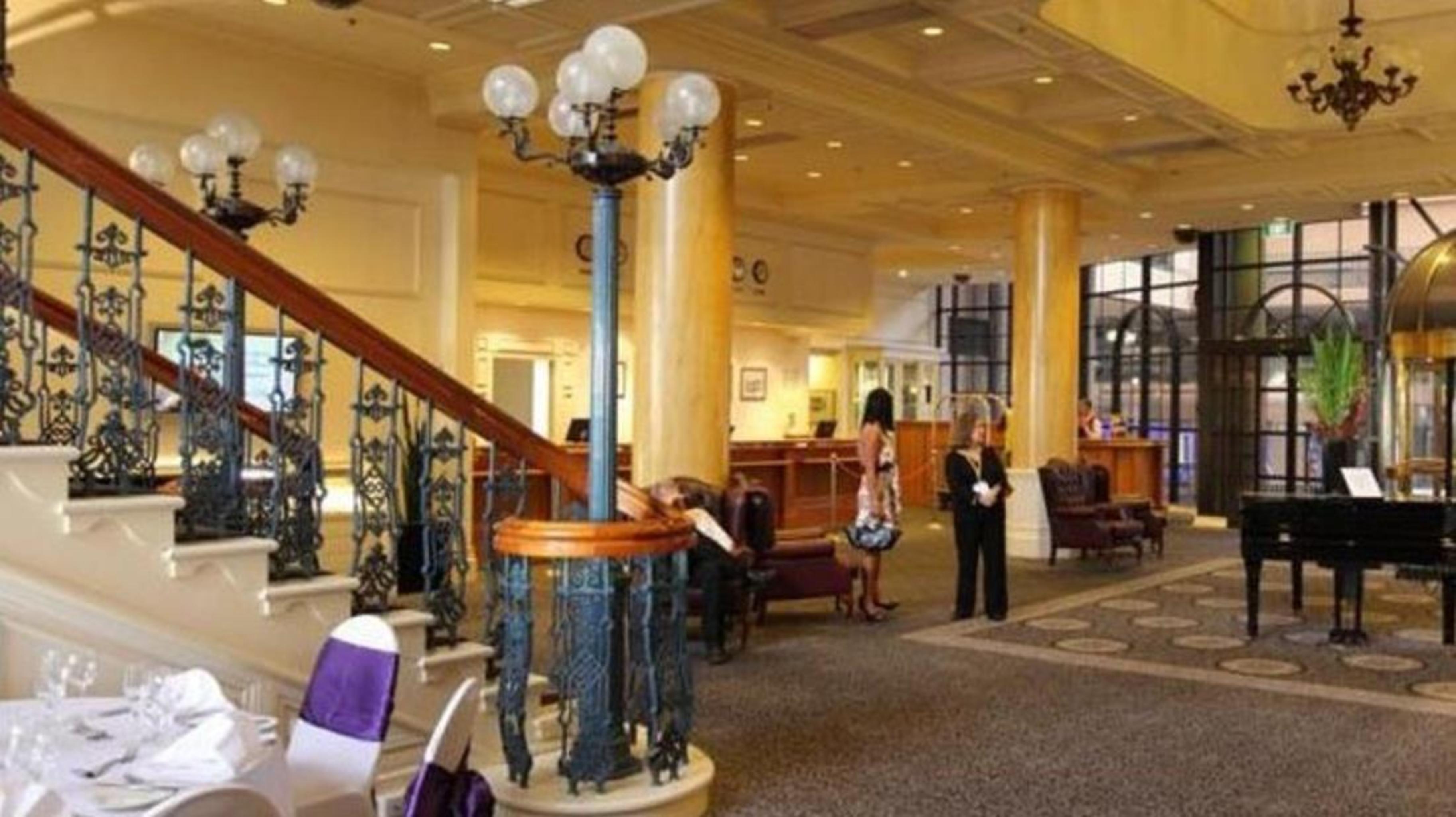 Stamford Grand Adelaide Hotel Glenelg Ngoại thất bức ảnh The photo shows the interior of a hotel lobby. It features a spacious area with a high ceiling and elegant decor. There is a staircase with a decorative railing leading to an upper level. In the foreground, some guests and staff are present, and ther