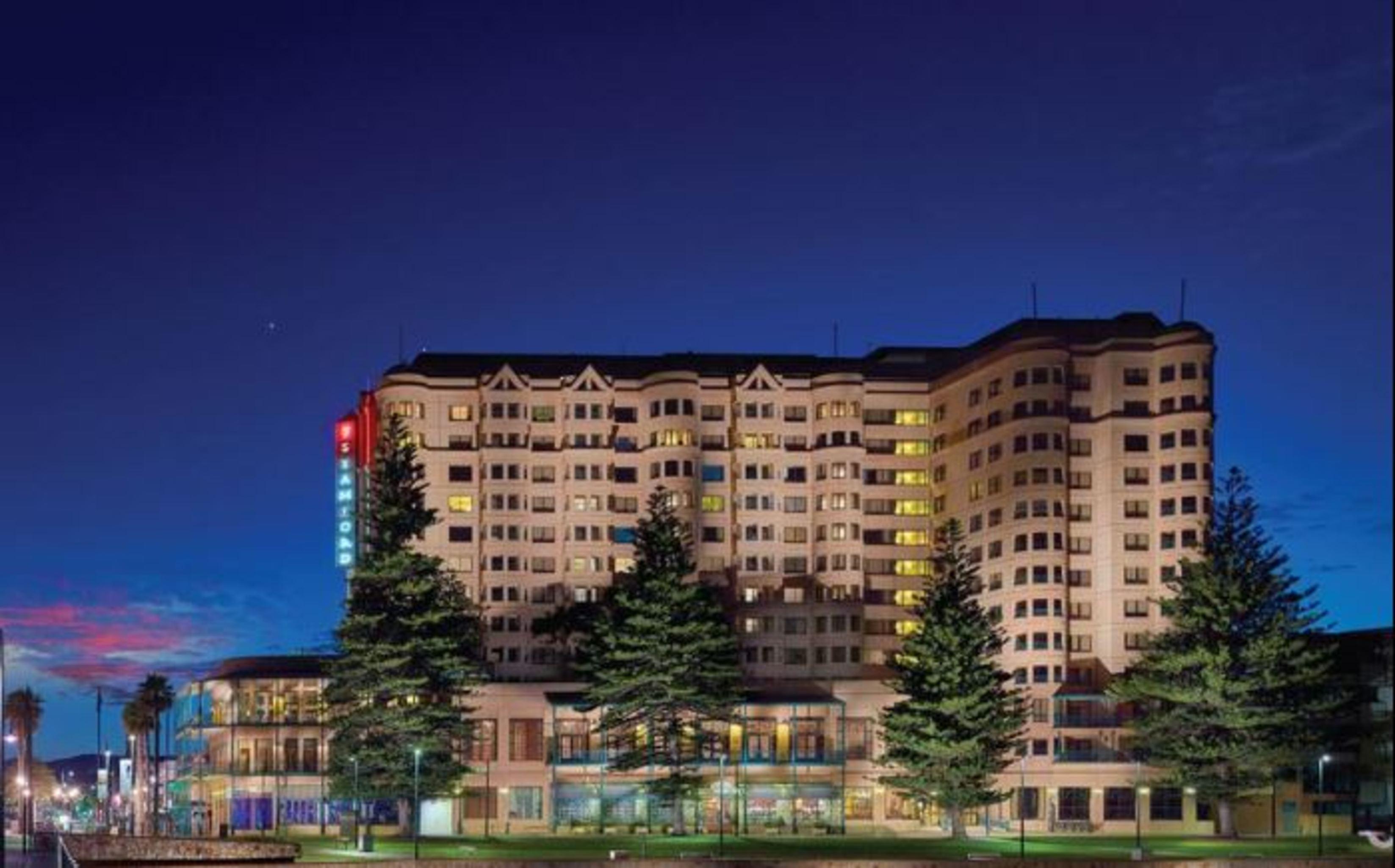 Stamford Grand Adelaide Hotel Glenelg Ngoại thất bức ảnh The photo displays a multi-story hotel building at dusk, illuminated by various lights. The architecture features a mix of modern and traditional design elements, with numerous windows showcasing a warm glow. Surrounding the hotel are several tall tr
