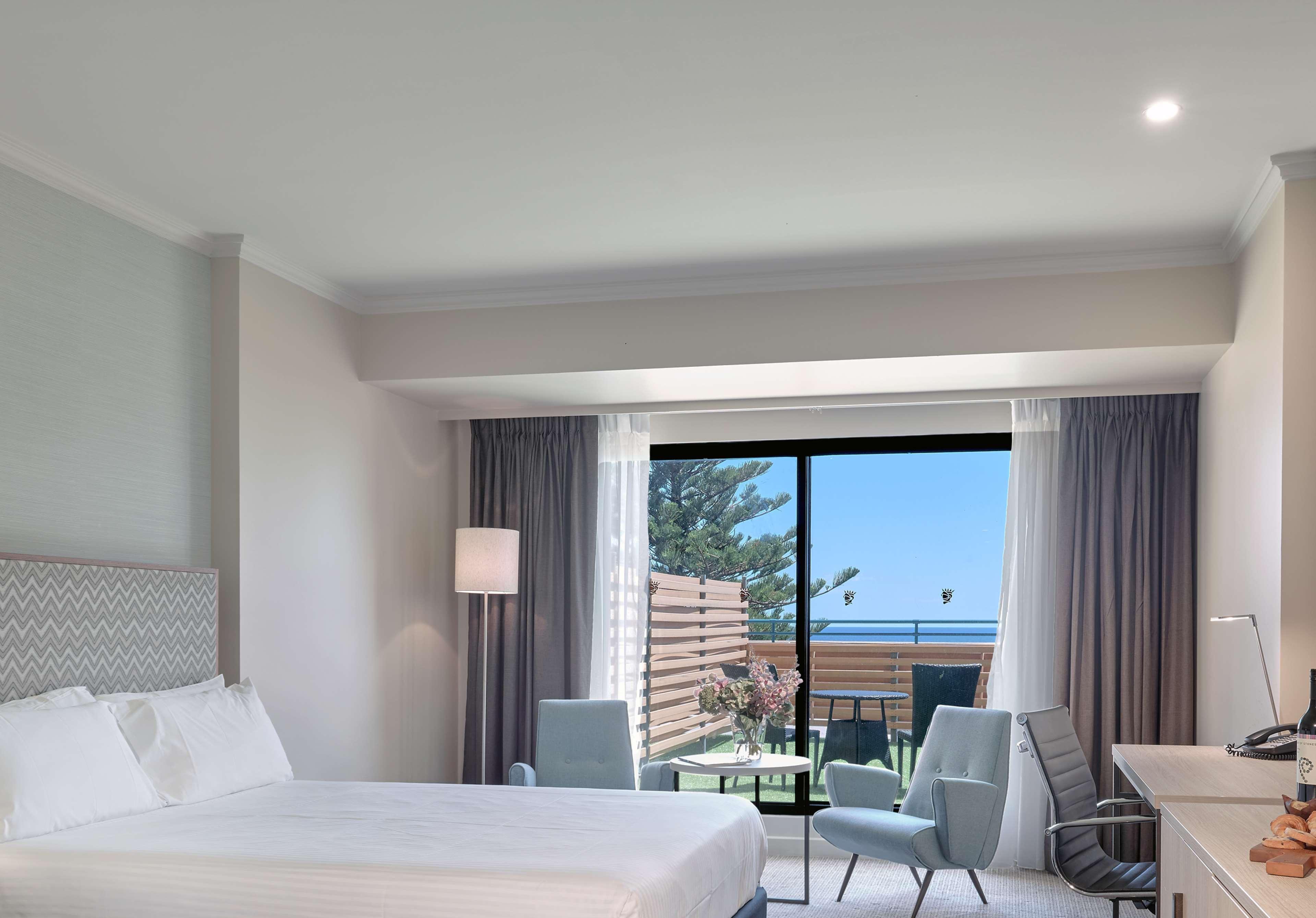 Stamford Grand Adelaide Hotel Glenelg Ngoại thất bức ảnh The photo shows a modern hotel room featuring a large bed with white bedding. There are two armchairs positioned near a window that offers a view of an outdoor area, possibly a balcony or patio with greenery. The room has light-colored walls and curt