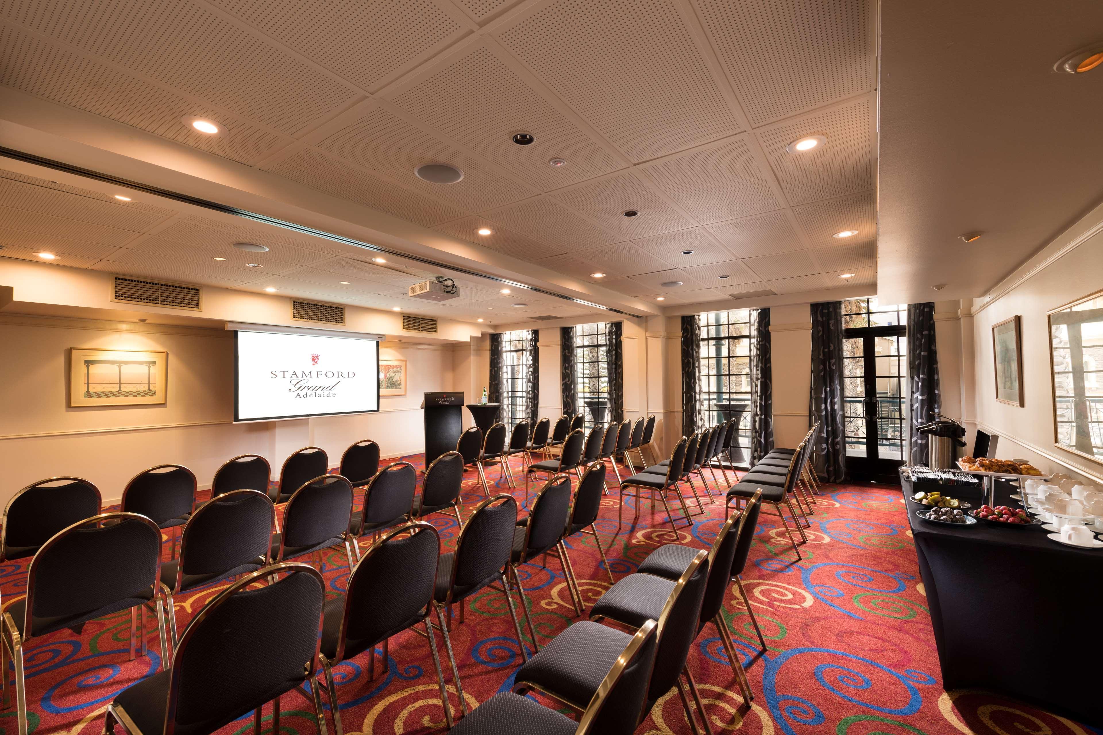 Stamford Grand Adelaide Hotel Glenelg Ngoại thất bức ảnh The photo shows a conference room set up for an event. There are rows of black chairs arranged in a semicircular formation facing a presentation screen at the front. The room features large windows that allow natural light to enter, and the flooring 