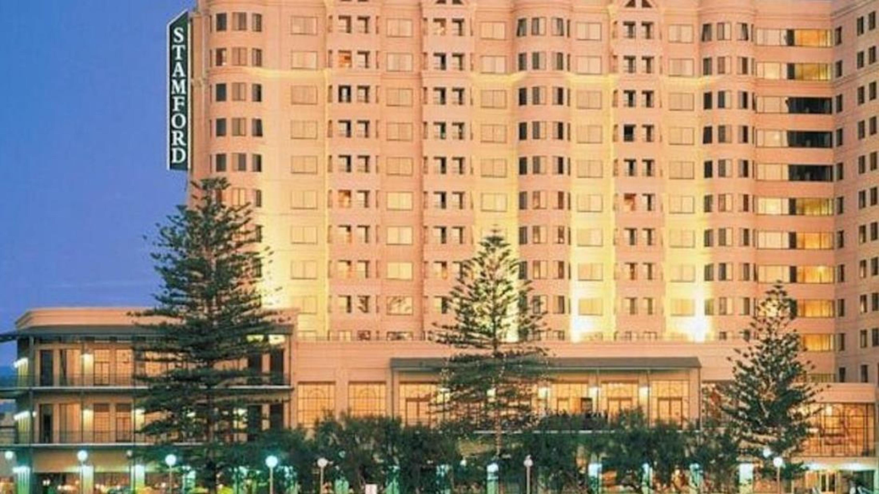 Stamford Grand Adelaide Hotel Glenelg Ngoại thất bức ảnh The photo shows a tall, modern building illuminated at night, likely a hotel, with the name "Stamford" prominently displayed on its side. Surrounding the building are several palm trees and landscaped areas, suggesting a tropical or coastal location.