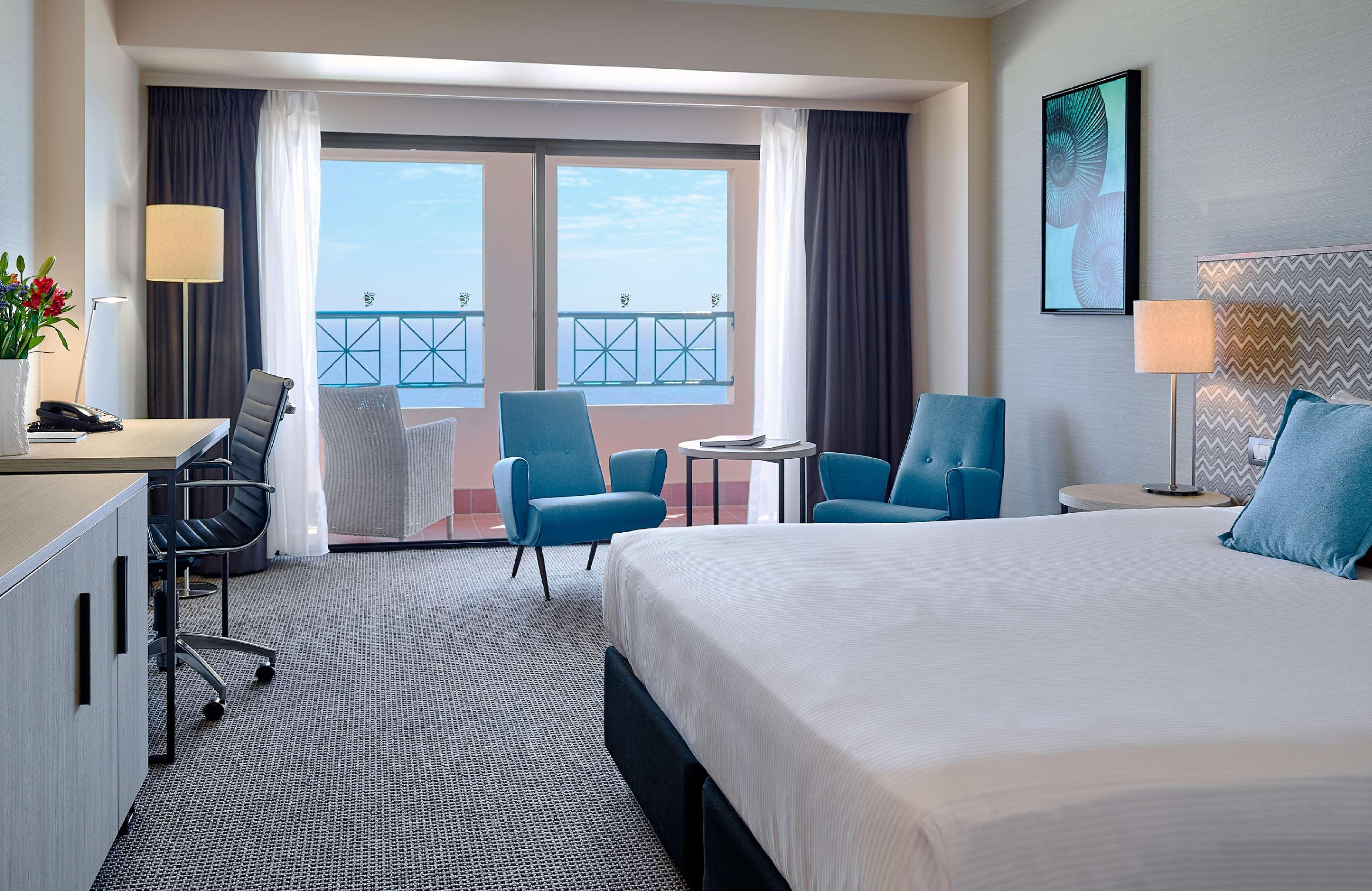 Stamford Grand Adelaide Hotel Glenelg Ngoại thất bức ảnh The image shows a hotel room featuring a large bed with a white duvet. There are two bright blue chairs and a small table near a large window that provides a view outside, possibly to a balcony. The room includes a workspace with a desk and an office