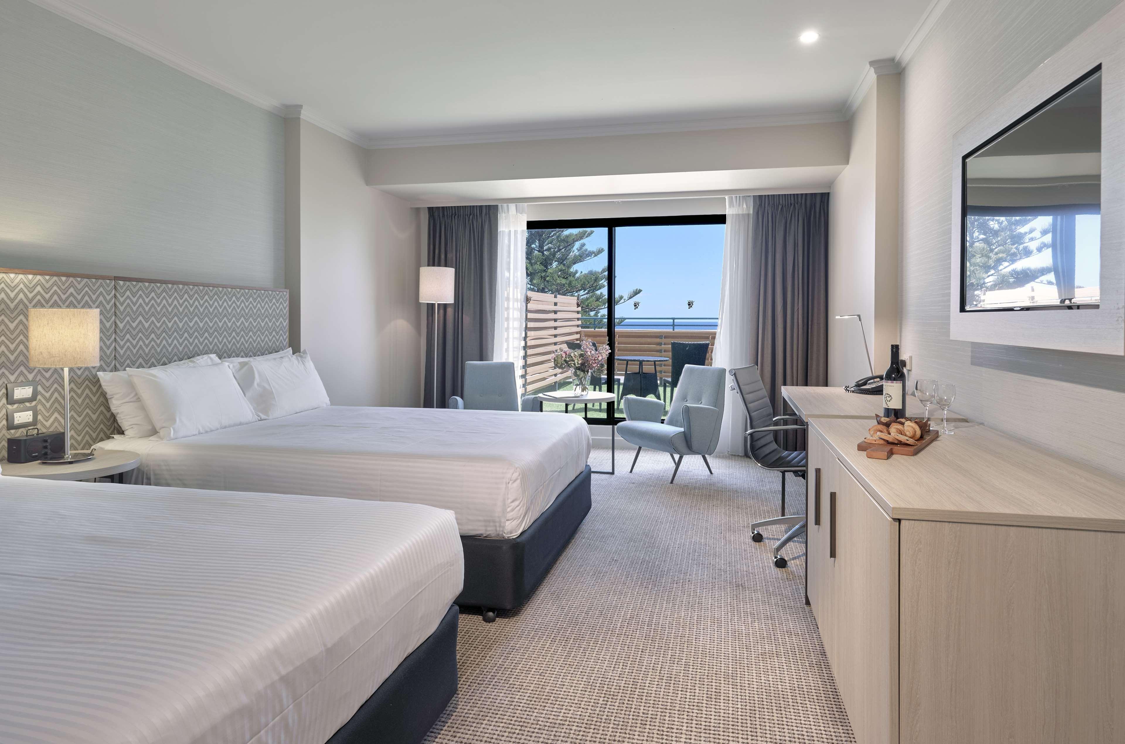 Stamford Grand Adelaide Hotel Glenelg Ngoại thất bức ảnh The photo shows a modern hotel room featuring two beds with white linens. There's a large window that provides natural light and a view of a balcony area. The room includes two chairs and a small table, creating a cozy sitting area. A television is m