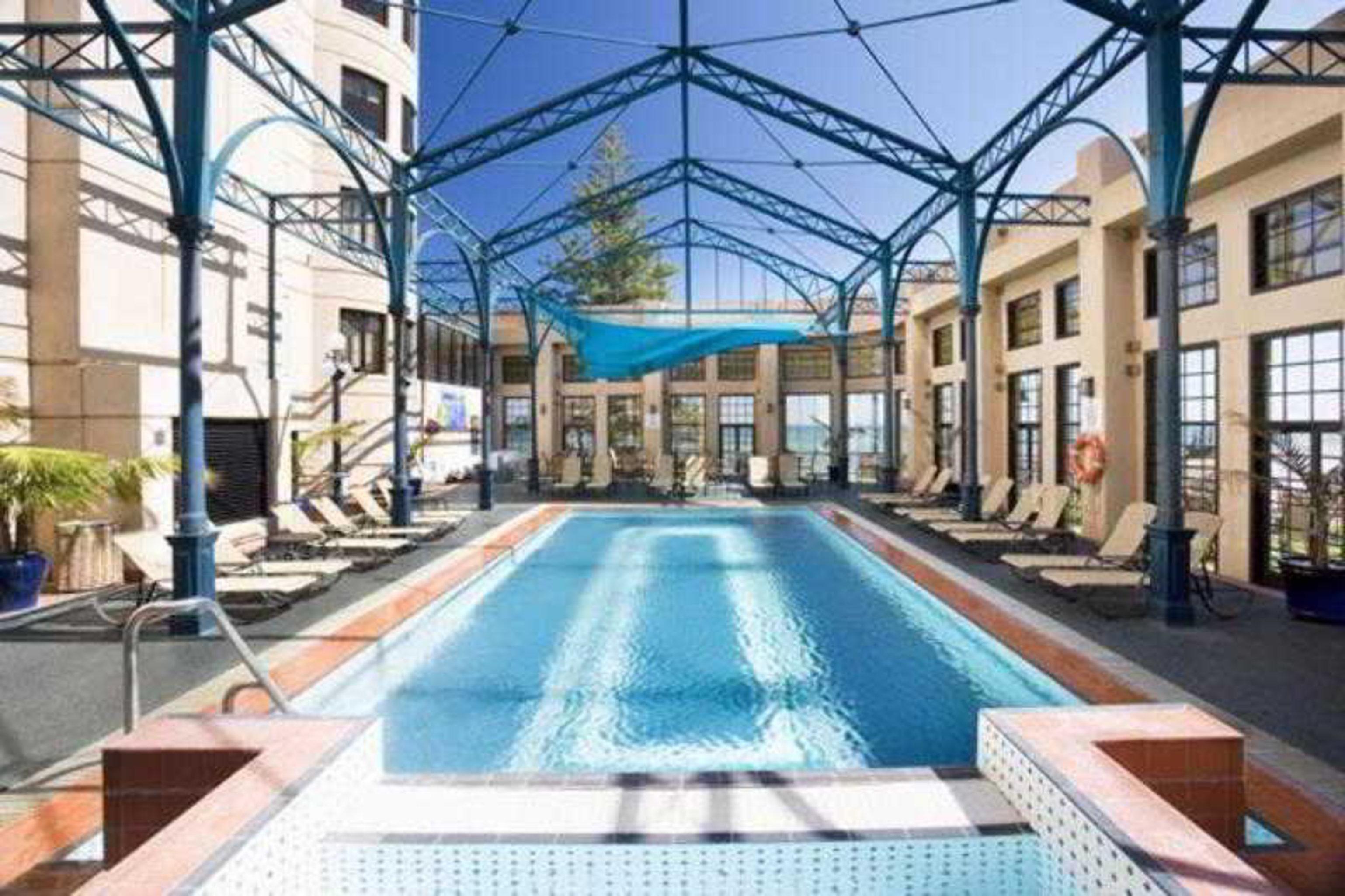 Stamford Grand Adelaide Hotel Glenelg Tiện nghi bức ảnh The photo shows an indoor swimming pool area. The pool is rectangular and features a blue cover above, supported by a metal frame. Surrounding the pool are several lounge chairs, and the space is well-lit with large windows allowing natural light to 