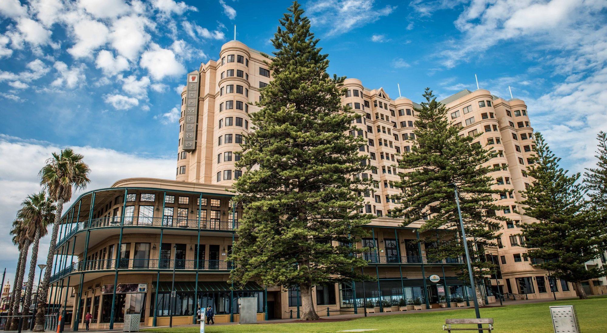 Stamford Grand Adelaide Hotel Glenelg Ngoại thất bức ảnh The image shows a large building with a distinctive architectural style, characterized by a tan or beige color. The structure appears to have multiple floors and rounded edges. In front of the building, there are several tall green trees, which add t