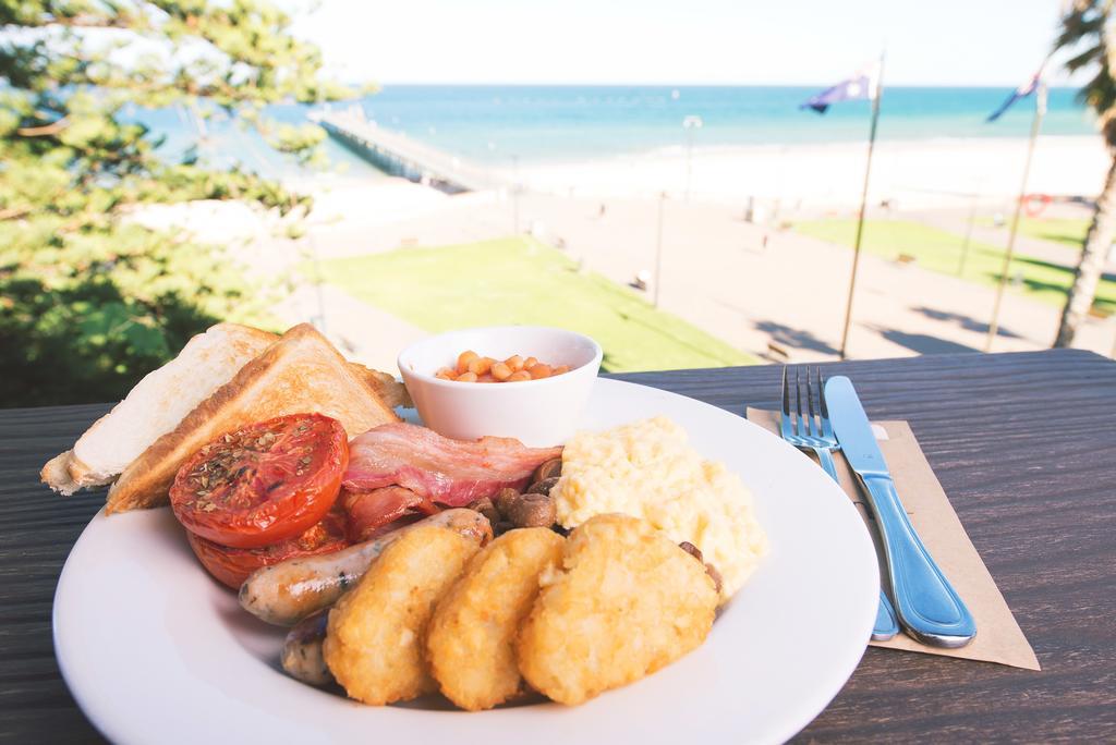 Stamford Grand Adelaide Hotel Glenelg Ngoại thất bức ảnh The photo shows a plate of breakfast food set against a beachside backdrop. The plate includes several items: crispy bacon, a grilled tomato, sausage links, golden hash browns, and a small serving of baked beans. There is also a piece of toast on the