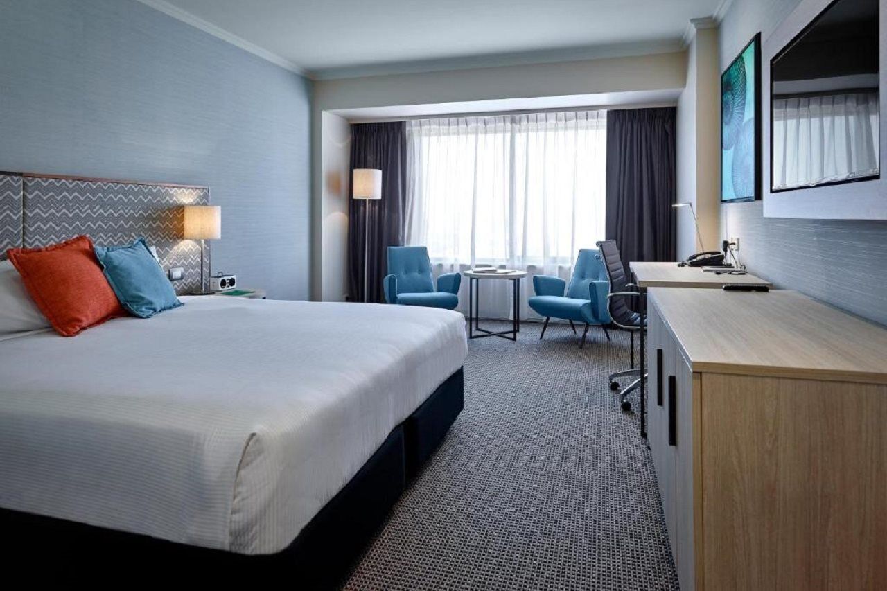 Stamford Grand Adelaide Hotel Glenelg Ngoại thất bức ảnh The photo shows a modern hotel room with a clean and minimalist design. In the center, there is a large, neatly made bed with white linens and two decorative pillows, one orange and one blue. To the right, there is a small wooden cabinet with a telev